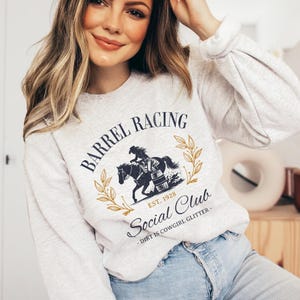 Pu&ograve; includere: Una felpa grigia con un design grafico di una donna a cavallo intorno a una botte. Il testo recita "Barrel Racing Est. 1928 Social Club - Dirt is Cowgirl Glitter - "