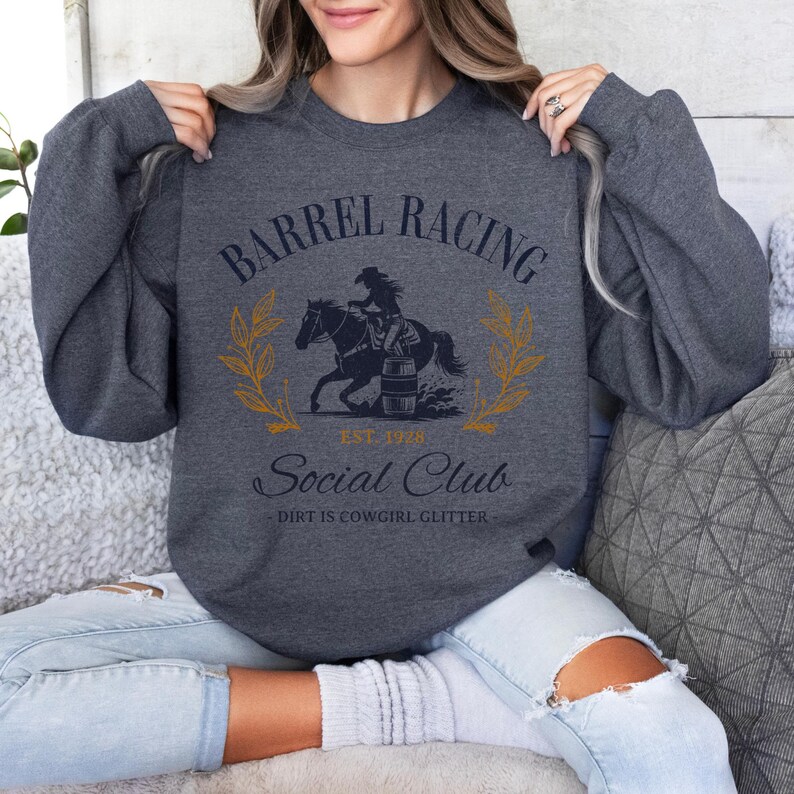 Pu&ograve; includere: Una felpa grigia scura con un design grafico in stile vintage che presenta una cowgirl a cavallo intorno a barili. Il testo recita "Barrel Racing Social Club Est. 1928 Dirt is Cowgirl Glitter".