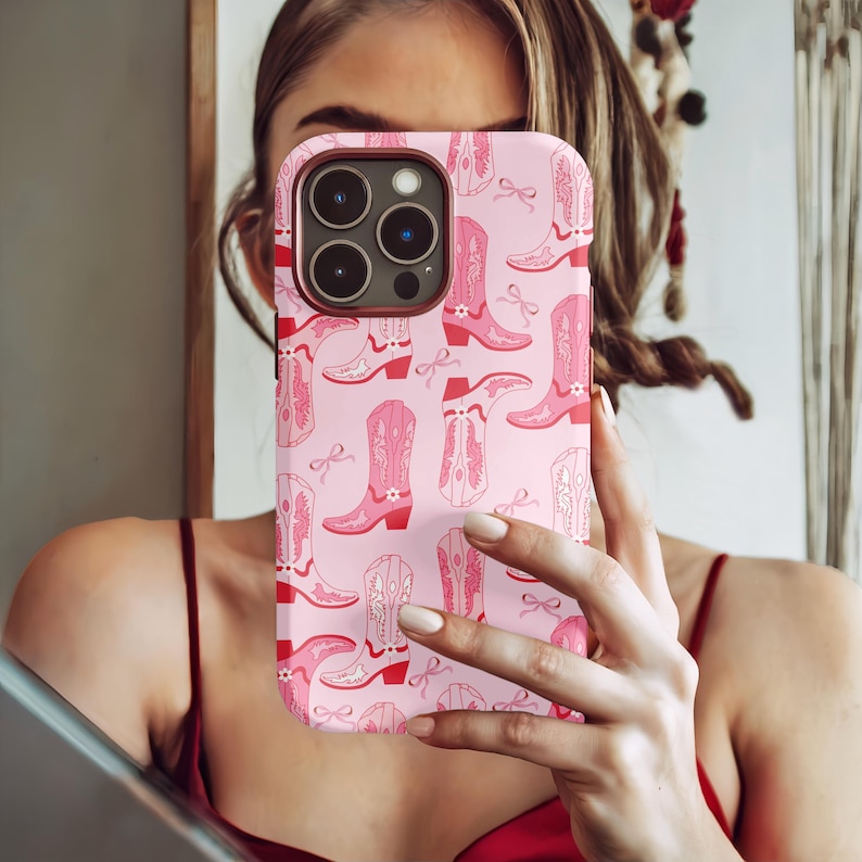 May include: Pink phone case with a repeating pattern of pink cowboy boots and bows. The case has a red border around the camera and edges. The boots have intricate details and are set against a light pink background.