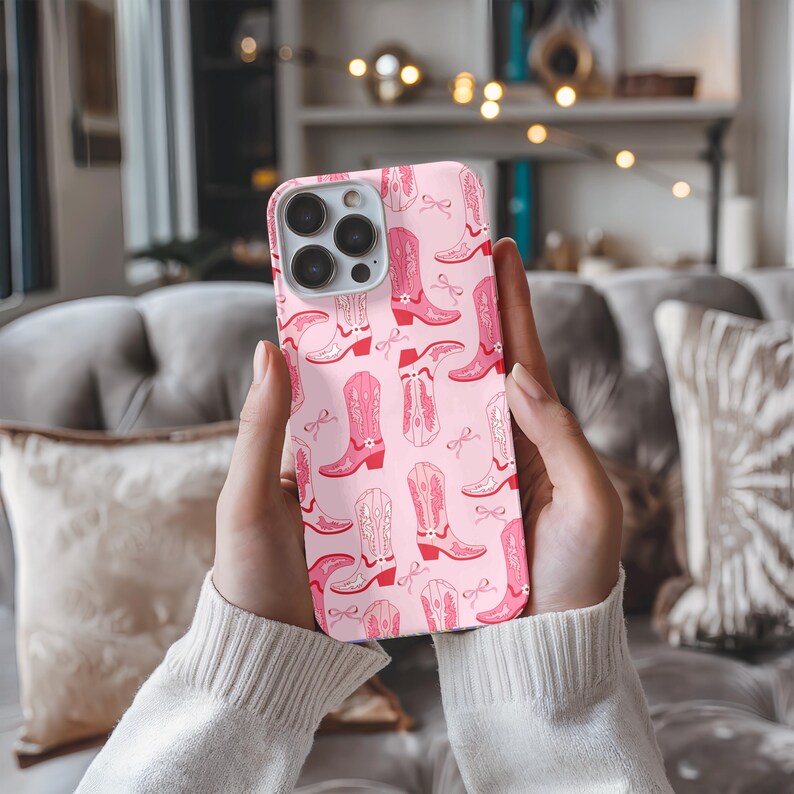 May include: A pink phone case featuring a repeating pattern of pink cowboy boots and bows. The boots vary in style and shade, with some having decorative stitching. The case is held in hands, with a soft background.