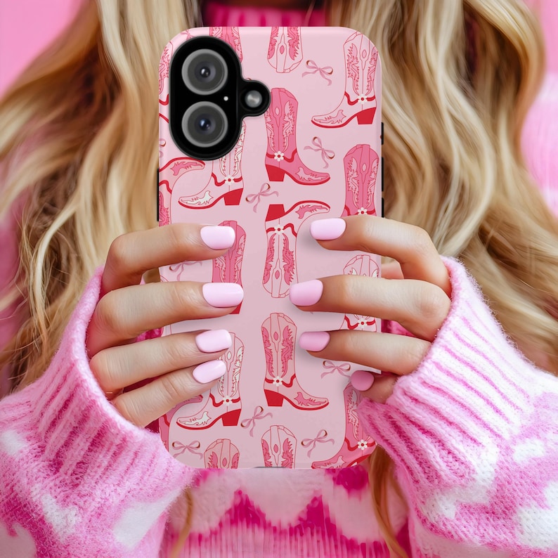 May include: A pink phone case featuring a repeating pattern of pink cowboy boots and bows. The case has a black camera lens cutout and is held in front of a pink sweater. The boots have red accents.