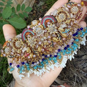 May include: Ornate gold-toned necklace with intricate floral and geometric designs. The necklace features colorful accents of blue, pink, and red gemstones, with dangling white beaded fringe. A detailed piece of jewelry.