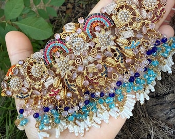 Op de afbeelding: Uitgebreide goudkleurige ketting met ingewikkelde bloemen- en geometrische patronen. Het heeft kleurrijke accenten van blauwe, roze en rode edelstenen, met bungelende witte kralenfranjes. Een gedetailleerd sieraad.