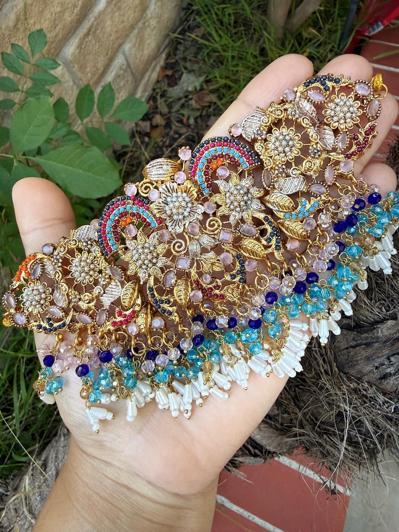 May include: Ornate gold-toned necklace with intricate floral and geometric designs. The necklace features colorful accents of blue, pink, and red gemstones, with dangling white beaded fringe. A detailed piece of jewelry.