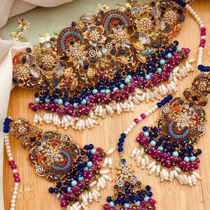 May include: An ornate gold-toned jewelry set featuring a necklace, earrings, and a headpiece. The set is adorned with colorful beads in shades of blue, purple, and white, along with intricate floral and geometric designs. The jewelry is displayed on a wooden surface.