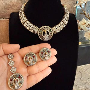 May include: An ornate gold-toned jewelry set featuring a necklace and earrings. The necklace has a wide band with white stones and a central pendant. The earrings and pendant have colorful stone accents.