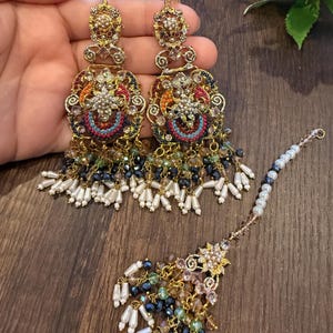 May include: Ornate gold-toned earrings and a matching headpiece. The earrings feature intricate filigree designs with colorful beadwork in red, blue, orange, and green, accented with dangling white and black beads. The headpiece has a floral design.