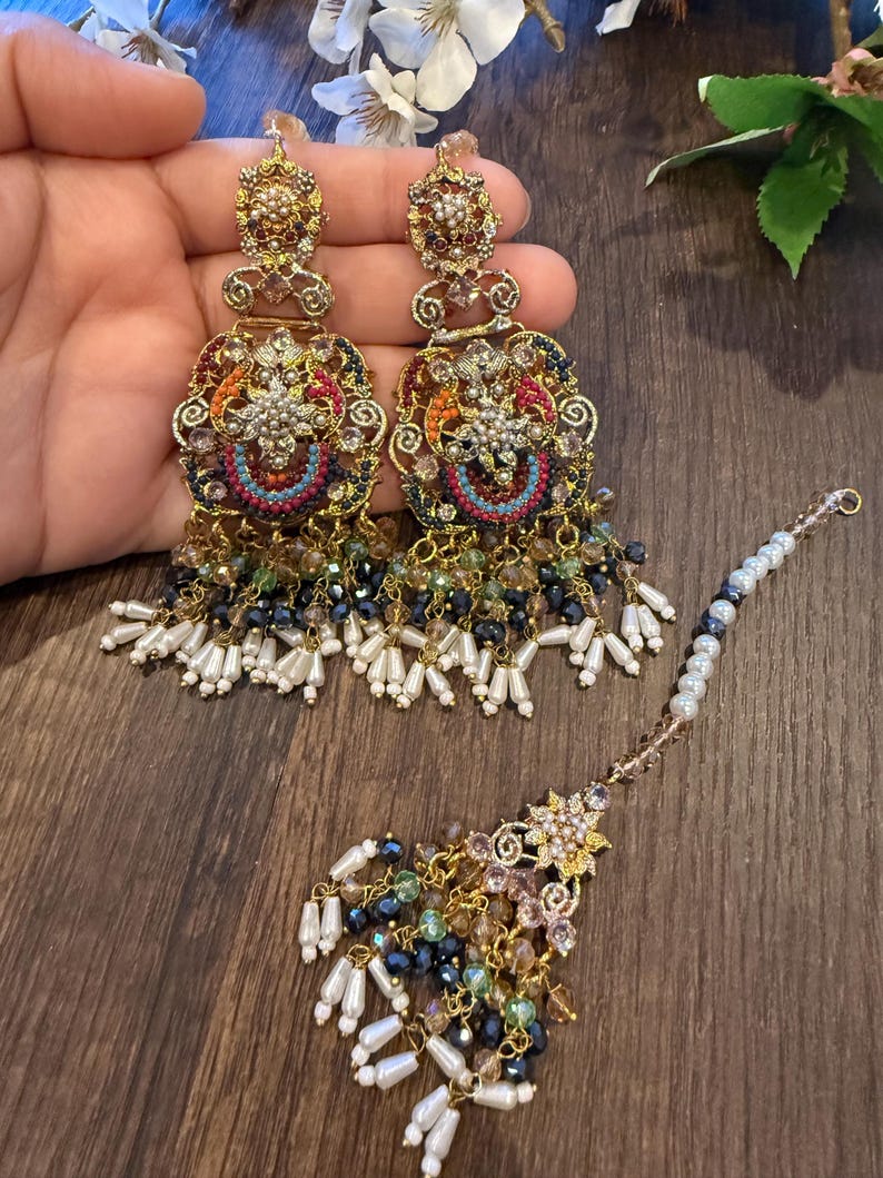 May include: Ornate gold-toned earrings and a matching headpiece. The earrings feature intricate filigree designs with colorful beadwork in red, blue, orange, and green, accented with dangling white and black beads. The headpiece has a floral design.