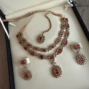 May include: A jewelry set featuring a double-strand necklace with rectangular pink stones surrounded by small clear crystals, a matching maang tikka with a single stone pendant, and two drop earrings with similar pink stones and crystal accents.