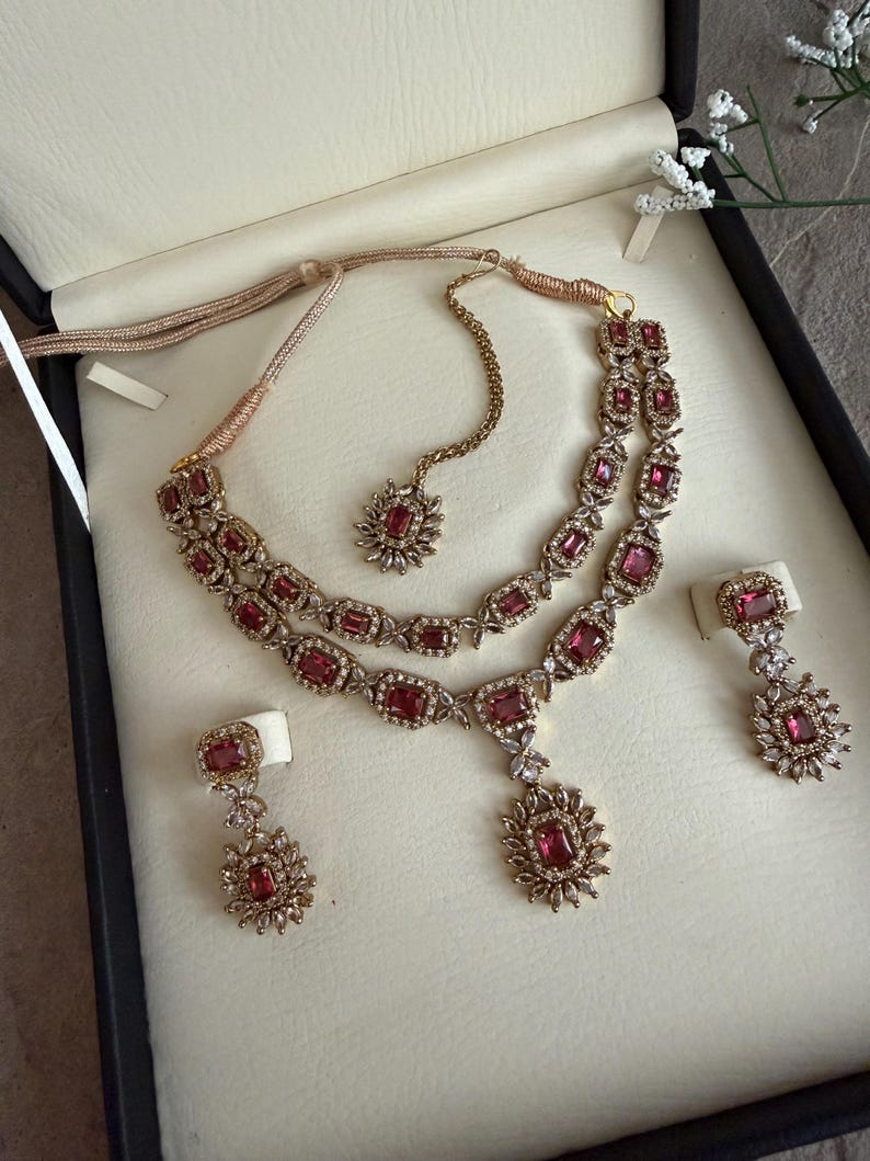 May include: A jewelry set featuring a double-strand necklace with rectangular pink stones surrounded by small clear crystals, a matching maang tikka with a single stone pendant, and two drop earrings with similar pink stones and crystal accents.