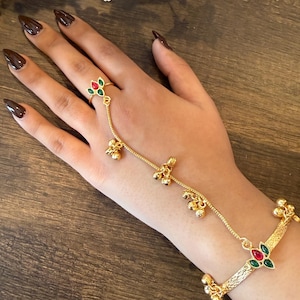 May include: Gold-toned hand chain bracelet with a ring, featuring red and green floral accents. The bracelet has dangling bell charms and a textured design. The ring is connected to the bracelet by a delicate chain.