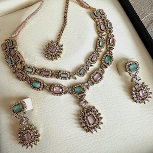 May include: A gold-toned jewelry set featuring a double-strand necklace with alternating rectangular pink and mint green stones, a matching maang tikka, and two drop earrings, all accented with small clear crystals in a floral-inspired setting.