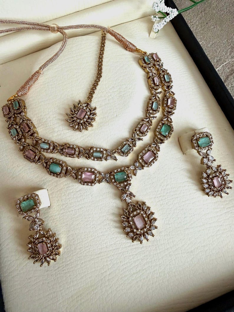 May include: A gold-toned jewelry set featuring a double-strand necklace with alternating rectangular pink and mint green stones, a matching maang tikka, and two drop earrings, all accented with small clear crystals in a floral-inspired setting.