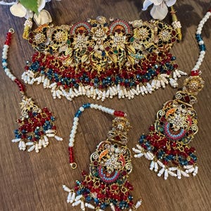 May include: An ornate gold-toned jewelry set featuring a choker necklace, earrings, and a headpiece. The set is adorned with red, blue, and white beads, along with intricate floral and geometric designs. The jewelry is displayed on a wooden surface.