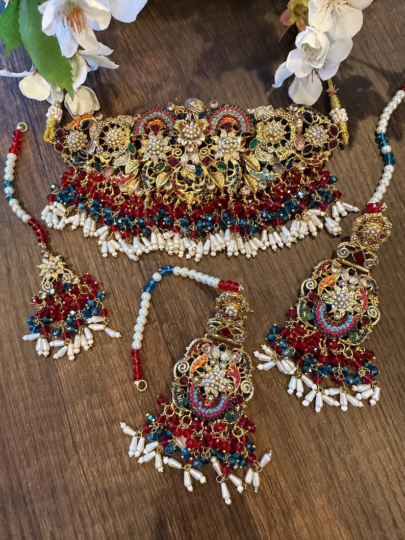 May include: An ornate gold-toned jewelry set featuring a choker necklace, earrings, and a headpiece. The set is adorned with red, blue, and white beads, along with intricate floral and geometric designs. The jewelry is displayed on a wooden surface.