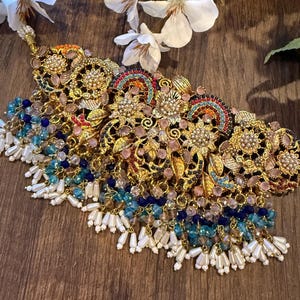 May include: An ornate gold-toned necklace with intricate floral and geometric designs. The necklace features colorful gemstones in shades of pink, blue, and orange, with dangling white pearl-like beads. White flowers are in the background.