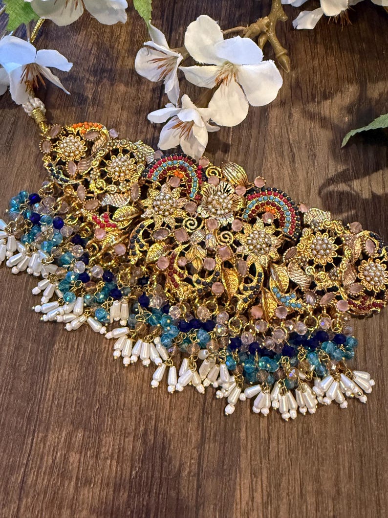 May include: An ornate gold-toned necklace with intricate floral and geometric designs. The necklace features colorful gemstones in shades of pink, blue, and orange, with dangling white pearl-like beads. White flowers are in the background.