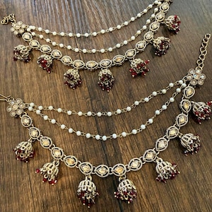 May include: A pair of gold-tone ear chains featuring multiple strands of small white beads. The base of each chain is adorned with metallic floral motifs, clear crystals, and dangling bell-shaped charms accented with deep red beads.