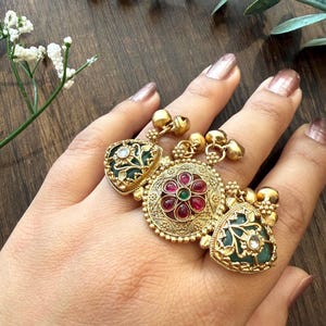 May include: A gold-toned statement ring with three ornate sections. The ring features emerald green and ruby red gemstones, intricate filigree, and small dangling bells. The ring is displayed on a hand with a neutral manicure.