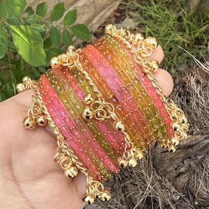 May include: A collection of colorful bangles in shades of pink, orange, and green. The bangles are adorned with gold-colored chains and small bell charms. The jewelry is held in a hand, with greenery in the background.