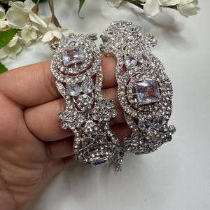 May include: Two silver-toned, ornate bangles encrusted with clear, sparkling rhinestones. Each bangle features a large, square-cut central stone and smaller, round stones. The design is intricate and detailed, with a floral-inspired pattern.