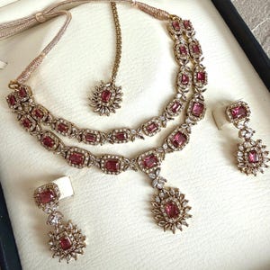 May include: A jewelry set featuring a double-strand necklace with rectangular pink stones and clear crystals, matching drop earrings, and a maang tikka. The pieces are set in a metallic frame with intricate crystal detailing and adjustable cords.