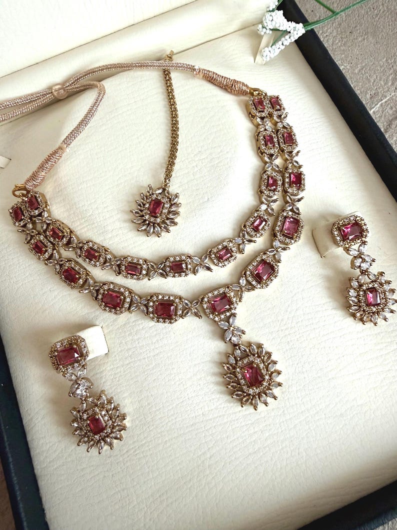 May include: A jewelry set featuring a double-strand necklace with rectangular pink stones and clear crystals, matching drop earrings, and a maang tikka. The pieces are set in a metallic frame with intricate crystal detailing and adjustable cords.