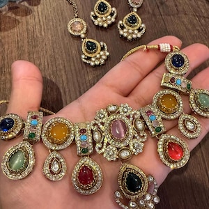 May include: A gold-toned jewelry set featuring a necklace, a pair of earrings, and a maang tikka. Each piece is adorned with multi-colored cabochon stones in shades of red, blue, yellow, and green, surrounded by small clear crystals and white beads.