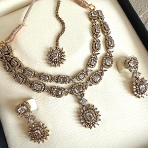 May include: A gold-toned jewelry set featuring a double-strand necklace with rectangular and floral-shaped clear crystals, a matching maang tikka with a central crystal pendant, and two dangling drop earrings with rectangular and floral crystal accents.