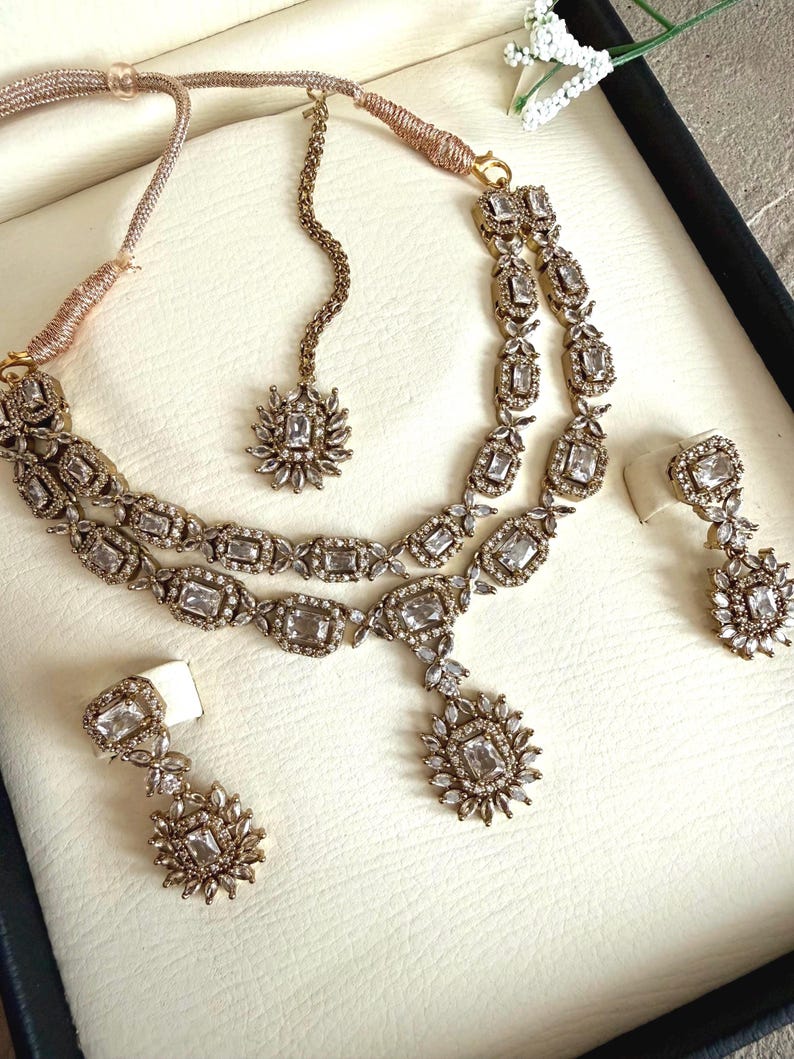 May include: A gold-toned jewelry set featuring a double-strand necklace with rectangular and floral-shaped clear crystals, a matching maang tikka with a central crystal pendant, and two dangling drop earrings with rectangular and floral crystal accents.