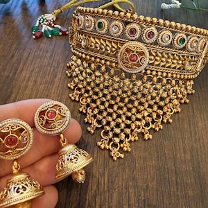 May include: A gold-toned choker necklace featuring intricate filigree patterns, embedded with small white, red, and green stones, and a dangling beaded fringe. Accompanied by matching jhumka-style earrings with circular tops and bell-shaped drops.