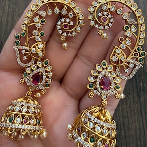 May include: A pair of gold-toned ear cuffs featuring an elongated, curved design adorned with clear, white stones, small green accents, and a central teardrop-shaped red stone. Each piece terminates in a bell-shaped jhumka pendant with pearls.