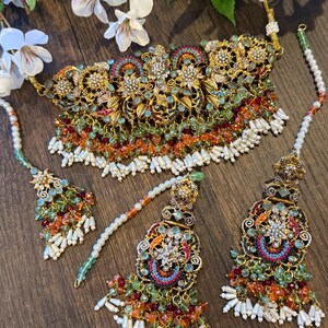 May include: An ornate gold-toned necklace and matching headpieces. The jewelry features intricate floral and geometric designs, with colorful bead accents in shades of green, orange, and red. White pearl-like beads hang from the bottom of the necklace and headpieces.