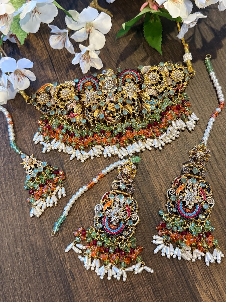 May include: An ornate gold-toned necklace and matching headpieces. The jewelry features intricate floral and geometric designs, with colorful bead accents in shades of green, orange, and red. White pearl-like beads hang from the bottom of the necklace and headpieces.