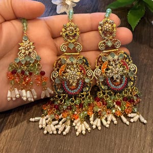 May include: Ornate gold-toned earrings with intricate detailing, featuring colorful beads in shades of orange, green, and red. The earrings have a chandelier design with dangling white pearl-like accents. A matching headpiece is also visible.