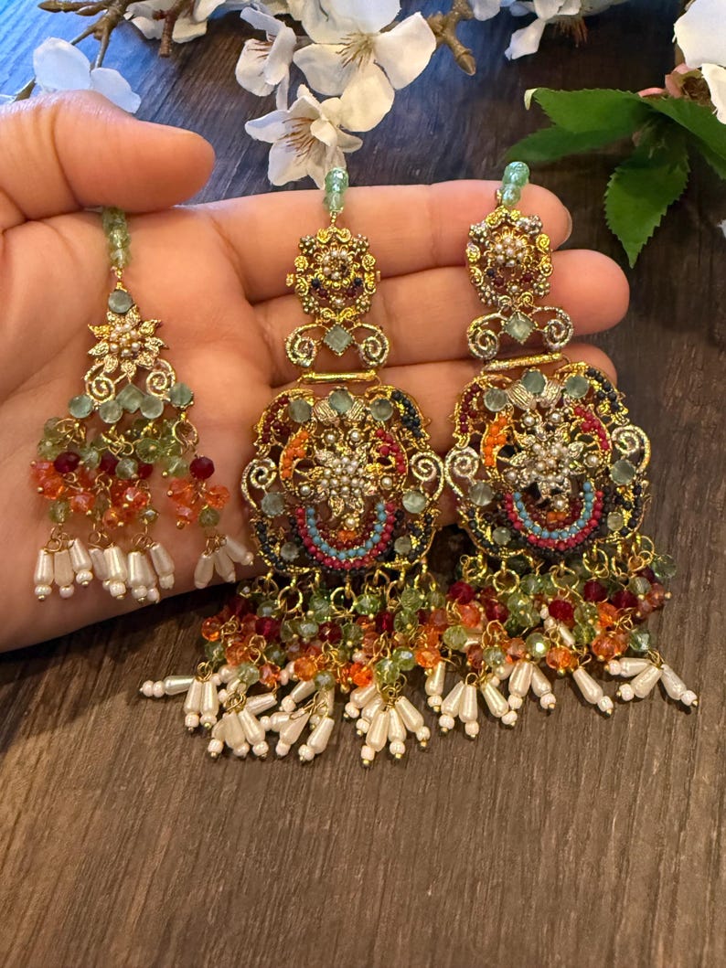 May include: Ornate gold-toned earrings with intricate detailing, featuring colorful beads in shades of orange, green, and red. The earrings have a chandelier design with dangling white pearl-like accents. A matching headpiece is also visible.
