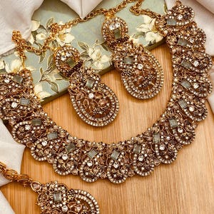 May include: A gold-toned jewelry set featuring a necklace, earrings, and a headpiece. The pieces are intricately designed with clear and pale green gemstones, surrounded by smaller, sparkling accents. The set is displayed on a wooden surface.