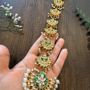 May include: A gold-toned maang tikka headpiece featuring a series of repeating floral-shaped motifs accented with green enamel and clear stones. The piece is finished with dangling white pearls and small white bead clusters along the edges.