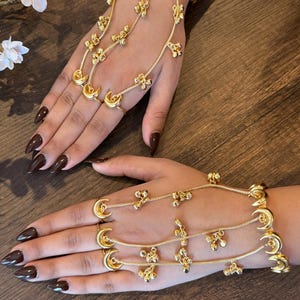 May include: Gold-toned hand jewelry featuring crescent moon and floral designs. The jewelry connects to rings and a bracelet, with delicate chains and decorative accents. The design is displayed on a wooden surface.
