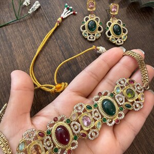May include: A gold-toned jewelry set consisting of a choker necklace and matching drop earrings. The pieces feature ornate metalwork frames set with oval-shaped dark green, deep red, and light pink stones, accented by small clear crystals.