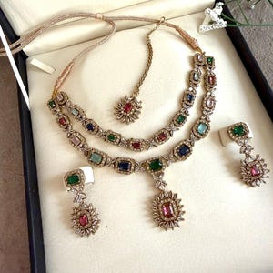 May include: A multi-piece jewelry set featuring a two-layered necklace, a maang tikka, and two drop earrings. Each piece is crafted with gold-toned metal, clear crystals, and rectangular-cut multicolored gemstones in shades of red, blue, and green.