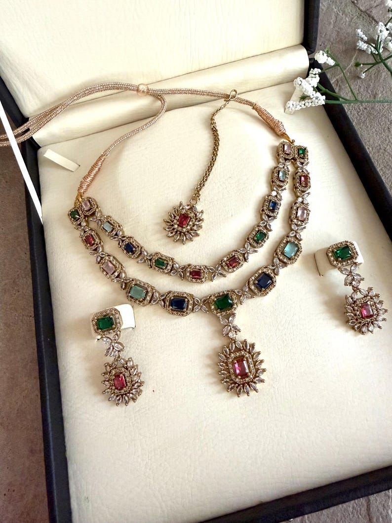 May include: A multi-piece jewelry set featuring a two-layered necklace, a maang tikka, and two drop earrings. Each piece is crafted with gold-toned metal, clear crystals, and rectangular-cut multicolored gemstones in shades of red, blue, and green.