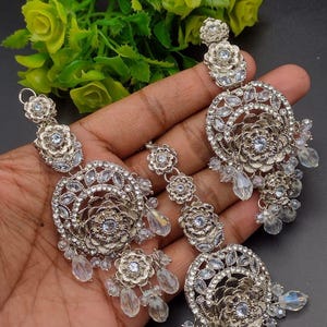 May include: A set of three silver-toned earrings featuring intricate floral motifs, clear crystal accents, and dangling teardrop-shaped beads. Each piece is constructed with layered circular patterns and small flower-shaped studs at the top.