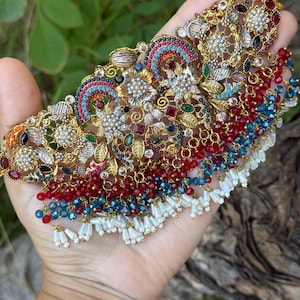 May include: Ornate gold-toned necklace with intricate floral and geometric designs. The necklace features colorful gemstones in red, blue, green, and clear, with dangling beaded accents and white tassels. The piece is held in a hand against a blurred green background.