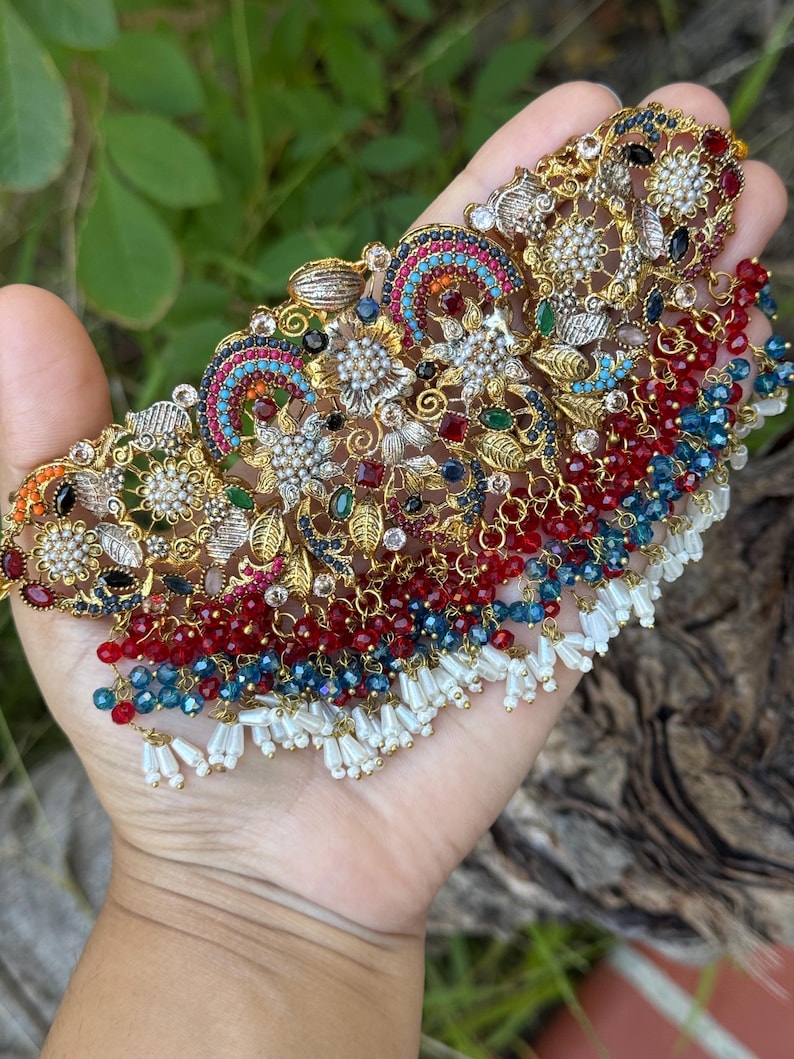 May include: Ornate gold-toned necklace with intricate floral and geometric designs. The necklace features colorful gemstones in red, blue, green, and clear, with dangling beaded accents and white tassels. The piece is held in a hand against a blurred green background.