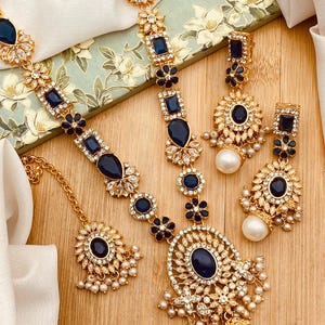 May include: A gold-toned jewelry set featuring a necklace, earrings, and a headpiece. The set is adorned with dark blue gemstones, clear crystals, and faux pearls. The necklace and earrings have a floral design.