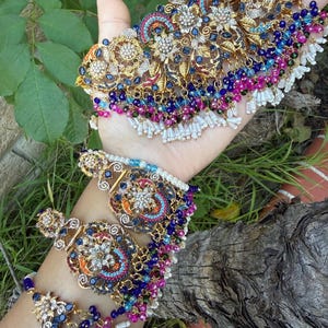 May include: An ornate gold-toned jewelry set featuring a necklace and bracelet. The pieces are adorned with intricate floral designs, sparkling gemstones in blue, pink, and white, and dangling beaded accents. The jewelry is displayed against a natural green background.