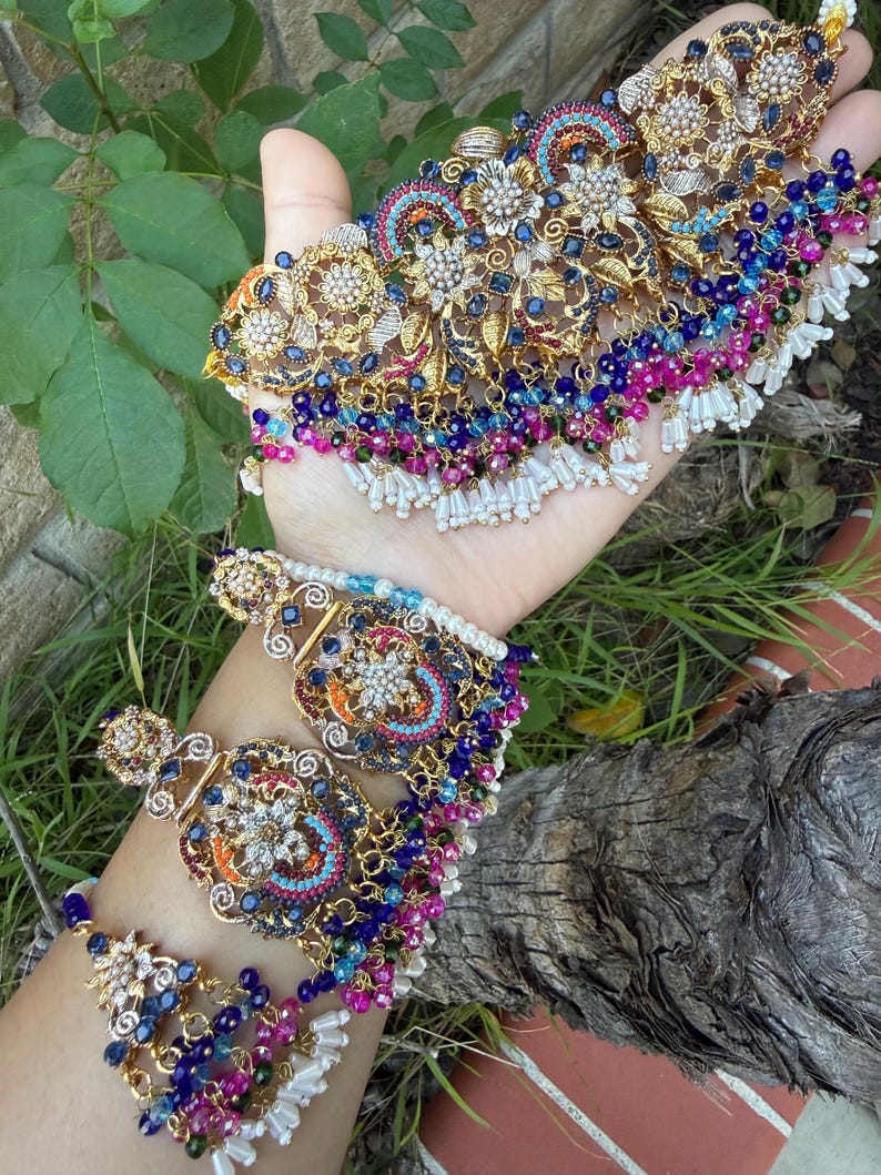 May include: An ornate gold-toned jewelry set featuring a necklace and bracelet. The pieces are adorned with intricate floral designs, sparkling gemstones in blue, pink, and white, and dangling beaded accents. The jewelry is displayed against a natural green background.