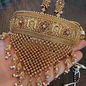 May include: A gold-toned choker necklace and matching earrings set featuring intricate metalwork, circular floral motifs with embedded red stones, and a wide, triangular-shaped fringe drop composed of small gold beads, white pearls, and red accents.