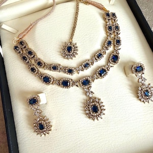 May include: A gold-toned jewelry set featuring a double-strand necklace with rectangular blue stones and clear crystals, a matching maang tikka with a central blue stone, and two drop earrings, each with a rectangular blue stone and crystal accents.